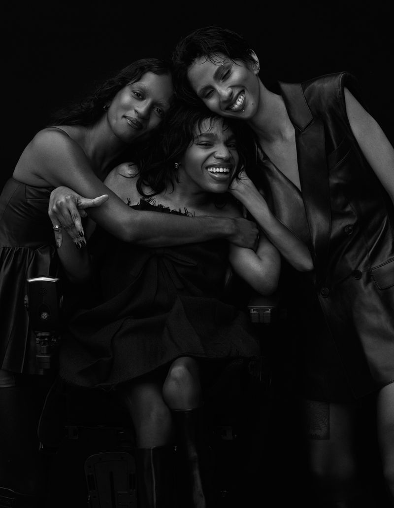 Three women in black and white embrace and smile in a photo studio setting, showcasing fashion and beauty in a close-up portrait.