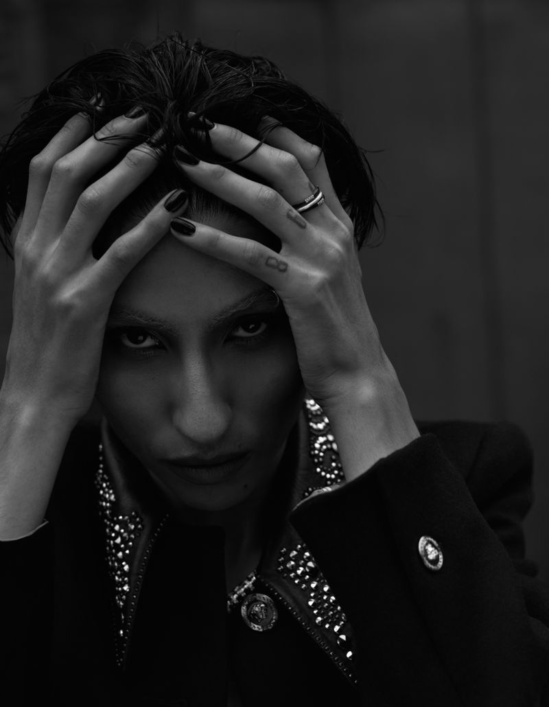 Portrait of a person with hands on their head in a black jacket, featuring dark hair and intense gaze in a stark black and white composition.