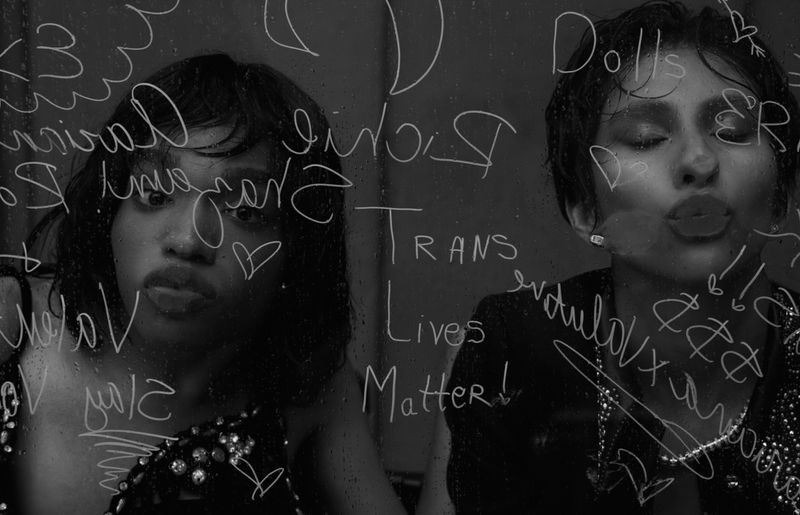 Black and white portrait of two adult women standing indoors behind a wet glass surface covered in handwritten words and symbols.