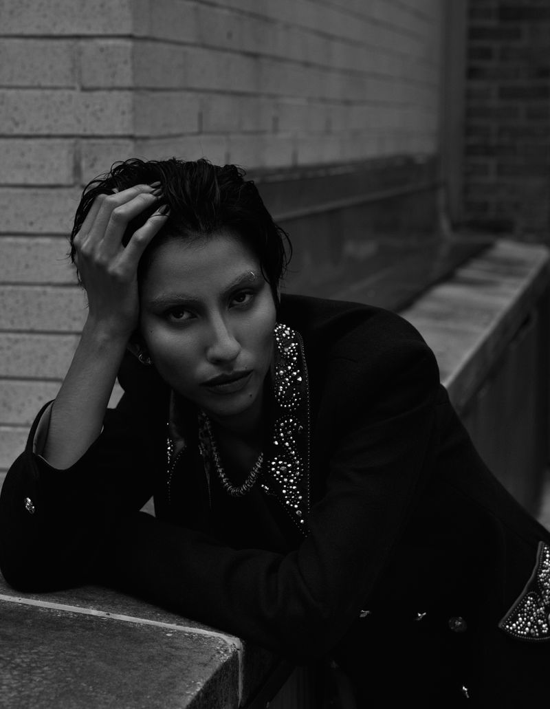 Monochrome portrait of a woman with short hair leaning against a ledge, wearing an embellished jacket in an urban setting.