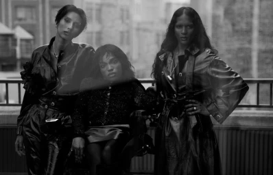 Monochrome editorial portrait of three models in dark coats and textured outerwear posing by a rooftop railing.