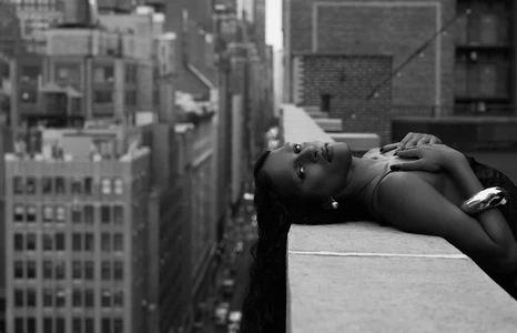 Black and white portrait of Richie Shazam reclining on an urban rooftop ledge overlooking city skyscrapers.