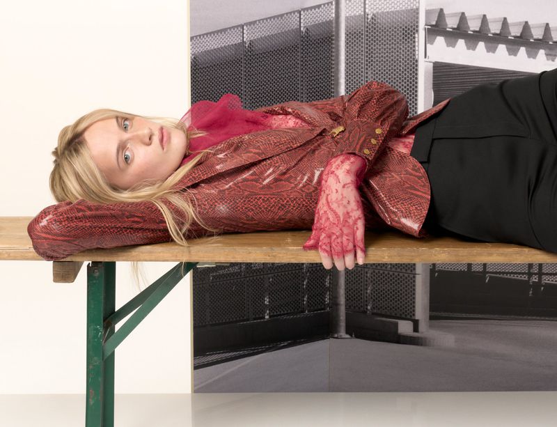 Young woman with long blonde hair lies on a wooden surface wearing a red snakeskin jacket and black pants in a fashion editorial.