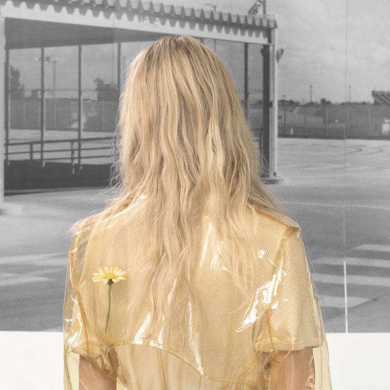 Blonde woman with back to camera wearing yellow jacket, with daisy flower pinned to her back, in front of modern building.