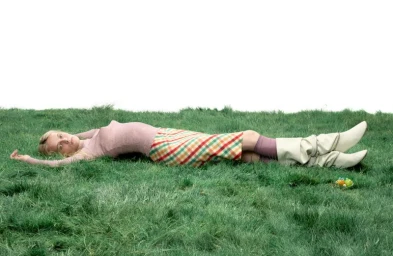 A blond woman lying horizontally on a patch of green grass against a white background, wearing a pink top, plaid skirt, and tall boots