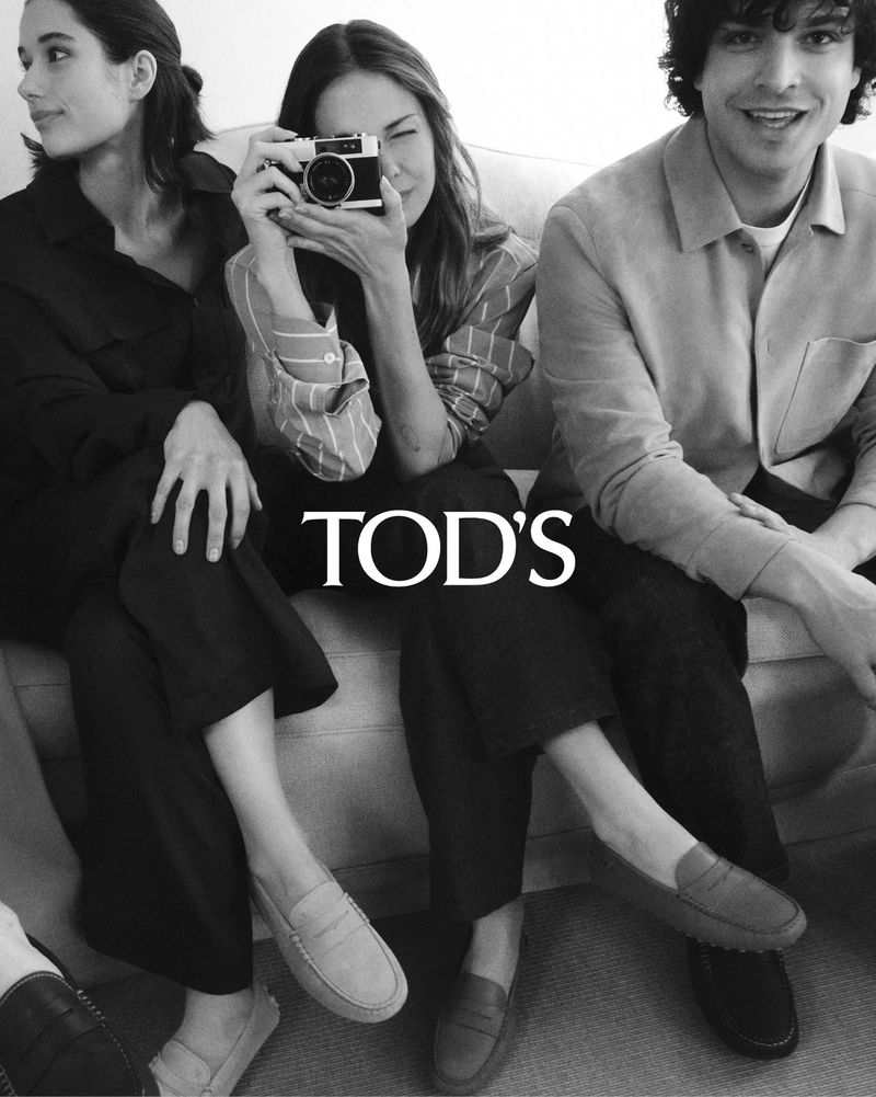 Fashion editorial shot of two women and a man posing with a camera and displaying Tod's loafers while seated in an indoor setting.