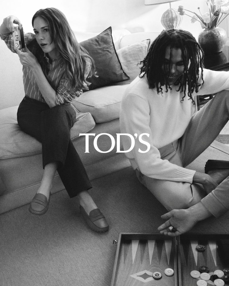 Black and white studio shot of a woman taking a photo of a man playing backgammon on the floor next to a couch. Tod's logo.