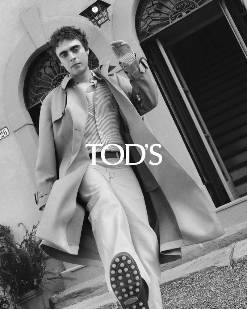 Monochrome shot of a male model in a trench coat and striped vest, with Tod's logo overlay, against an architectural backdrop.