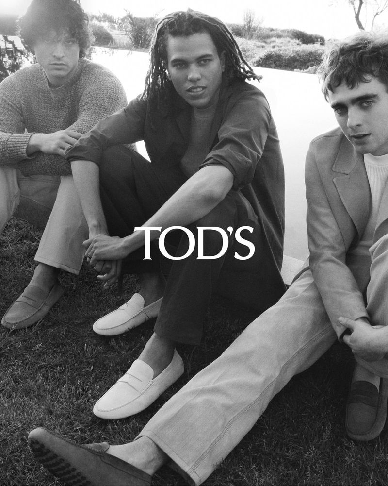 Three young adult men in a black-and-white fashion editorial sit on grass near water, showcasing TOD'S loafers and casual wear.