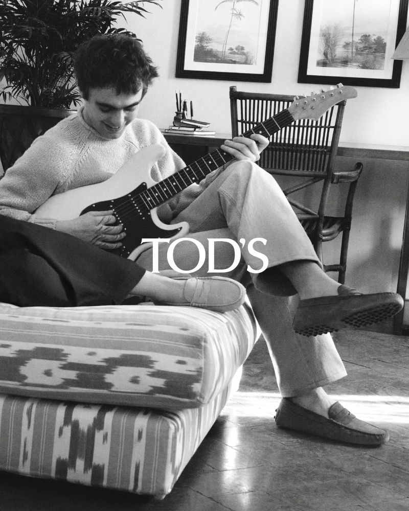 Black and white image of a casually dressed man playing an electric guitar while sitting on an ottoman in a comfortable room.