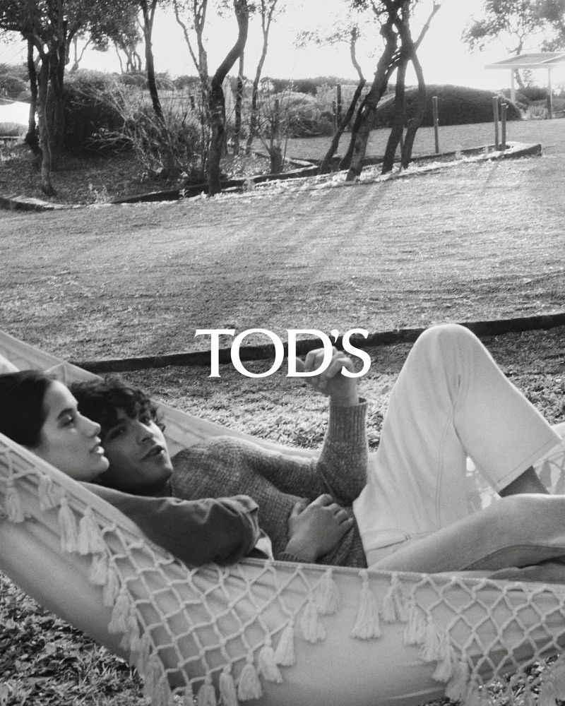 Monochrome portrait of a couple lounging in a rope hammock outdoors surrounded by grass and trees with visible brand logo.