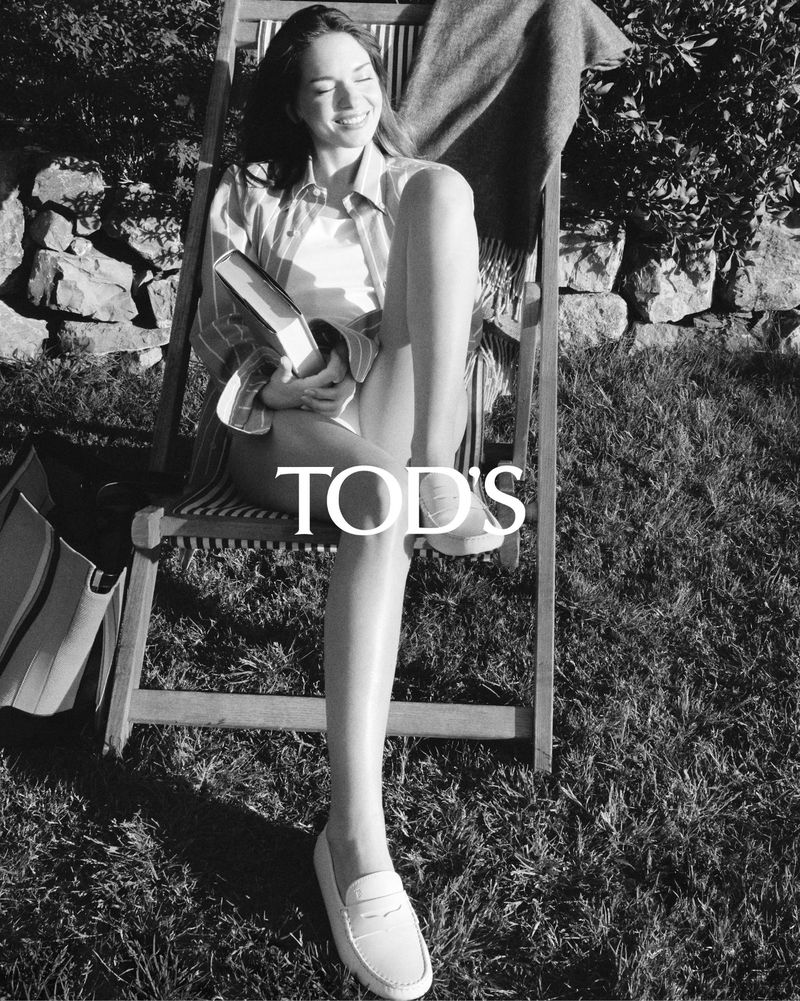 A smiling woman relaxes in a chair on the grass, wearing casual clothing and white loafers, with a book in her hands, outdoors.