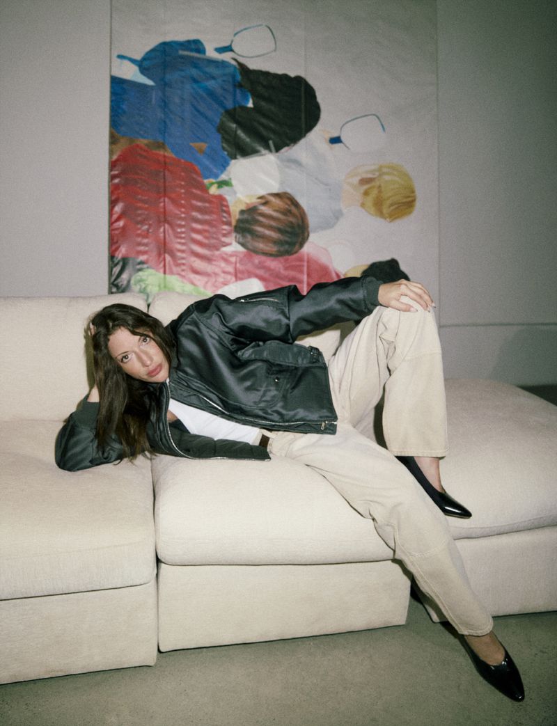 A young woman is lying on a couch wearing a black jacket, white top, and beige pants, with a modern art painting in the background.