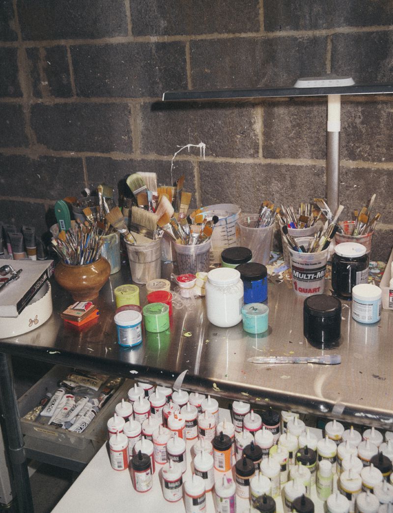Assorted paintbrushes and paint supplies arranged on a metal table against a brick wall in an art studio setting with studio lighting.