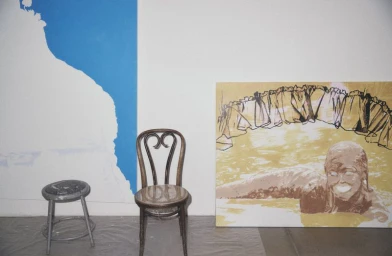 Artist studio with a bentwood chair, a round stool, and a large figurative painting leaning against a white and blue wall.