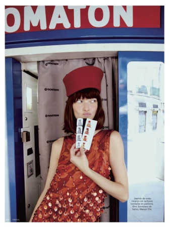 Attractive brunette in an orange dress and red hat holding photo booth strip while standing in a photo booth with red and blue trim.