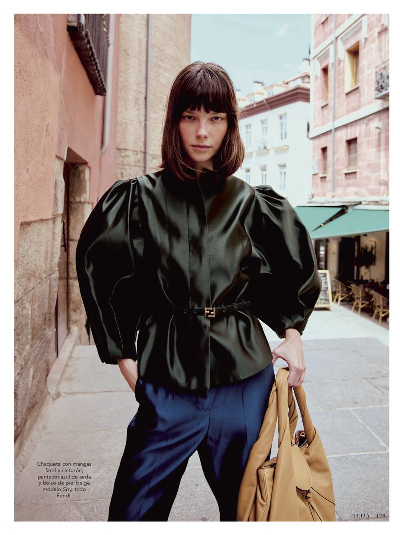 A woman in an emerald green jacket and blue pants stands on a city street with stone walls and buildings in the background.