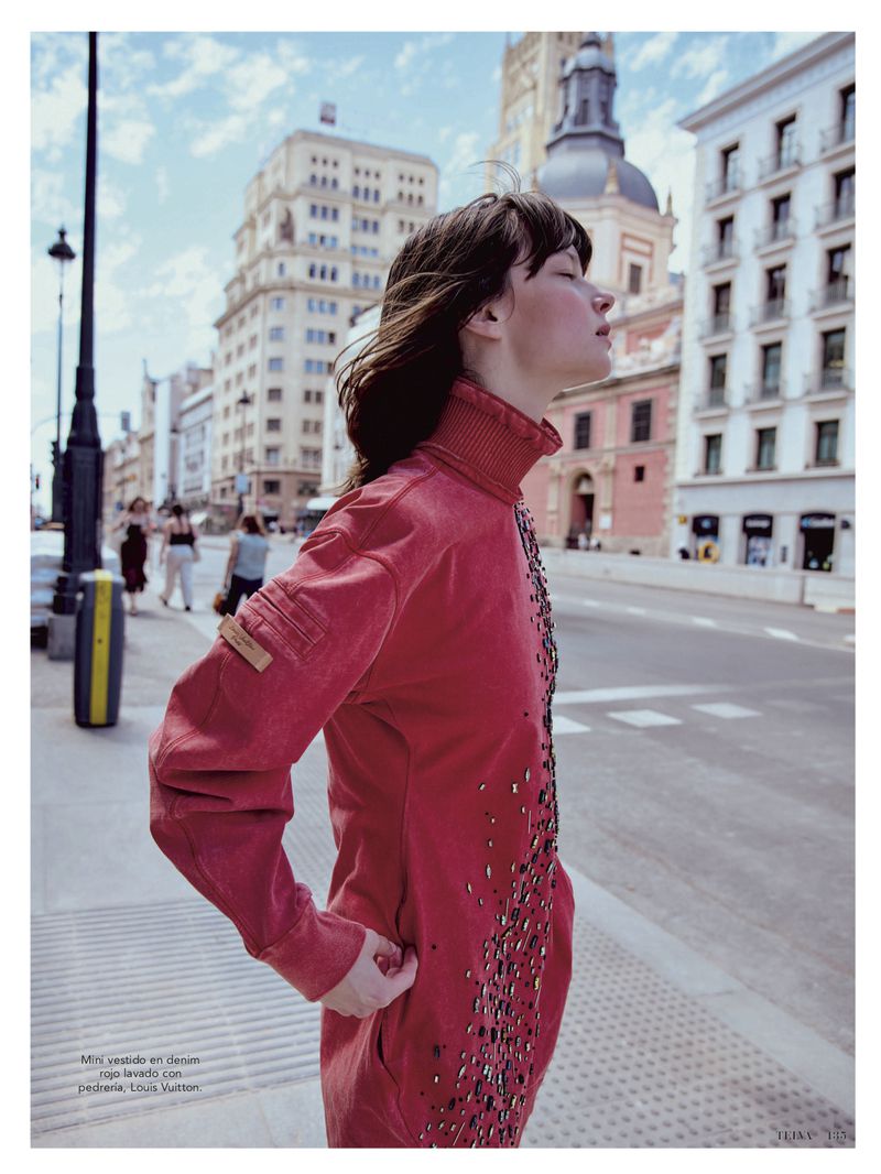 Woman in red jumpsuit stands on city sidewalk with historic buildings in the background, enjoying the sunlight in urban fashion editorial.