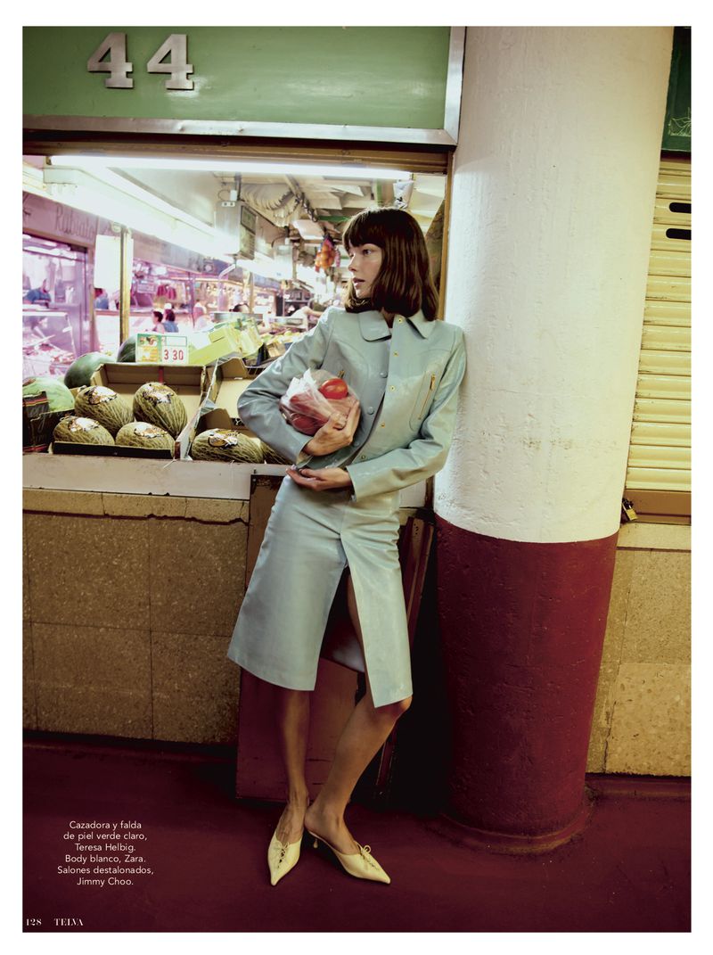 A woman in a light blue skirt suit leans against a column in a market setting, holding produce in a plastic bag.