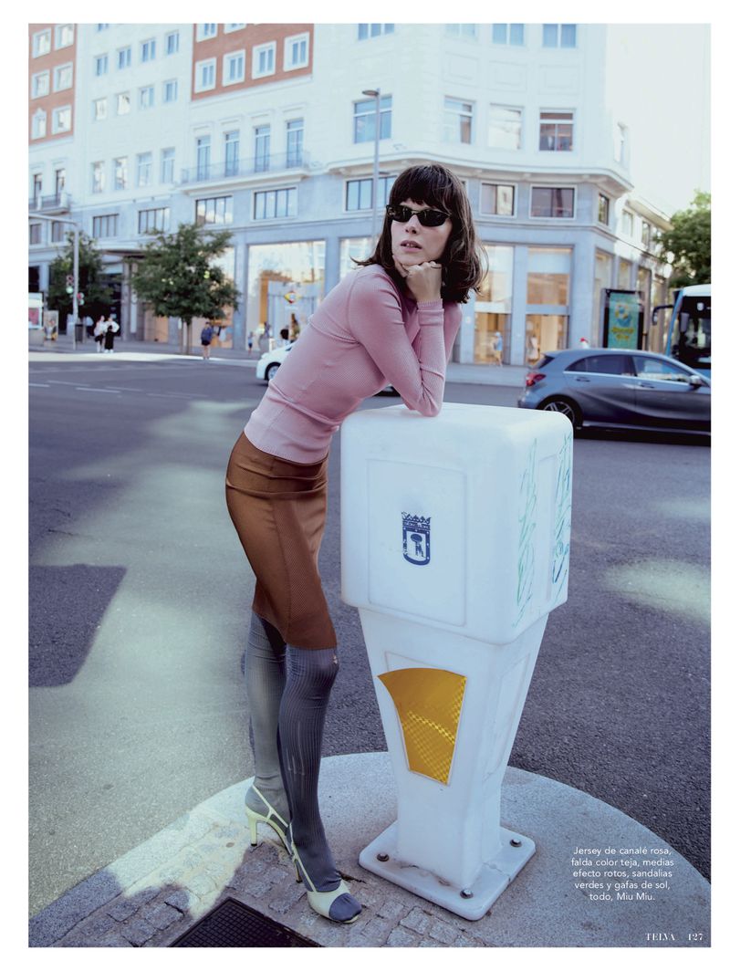 A woman in a pink sweater and brown skirt poses on a city street next to a white traffic barrier against a backdrop of urban buildings.