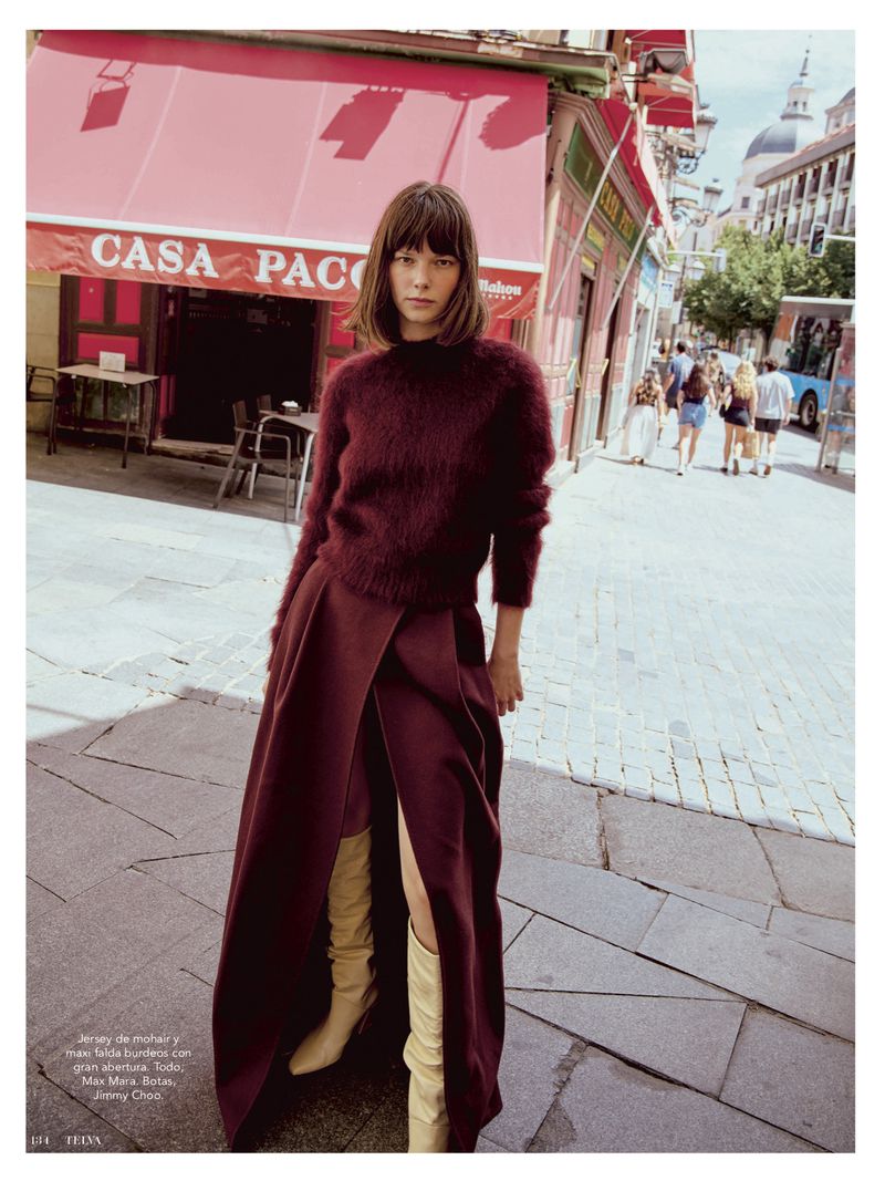 A woman stands on a sidewalk in a maroon sweater and skirt with tall beige boots, posing in front of a cafe in an urban setting.