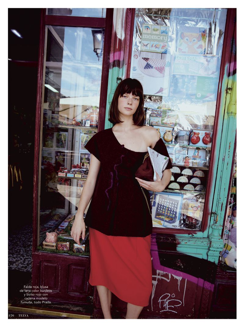Brunette woman in a black top and red skirt stands in front of a newsstand with magazines, toys, and snacks, natural lighting.