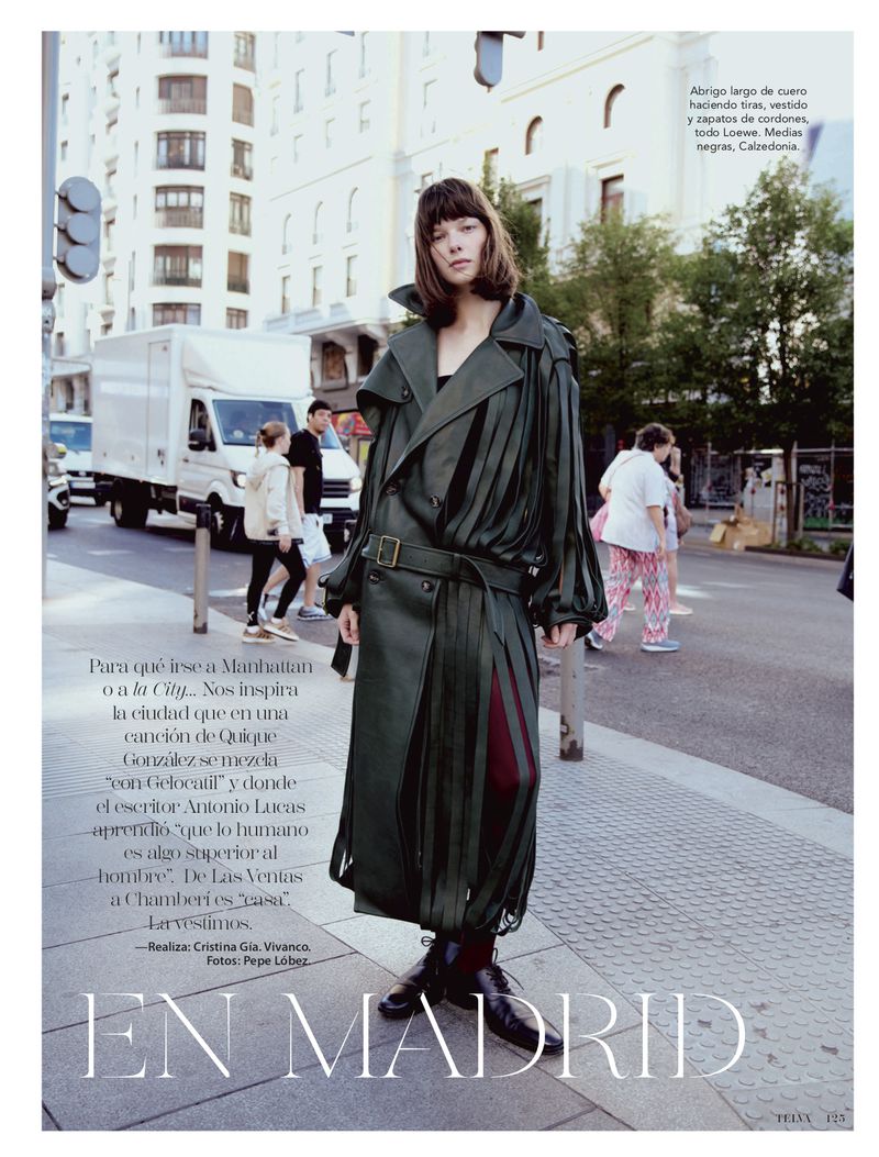 A woman in a long, olive-green leather coat stands on a city street in an editorial fashion photo. Pedestrians and a truck are in the background.