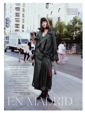 A woman in a long, olive-green leather coat stands on a city street in an editorial fashion photo. Pedestrians and a truck are in the background.