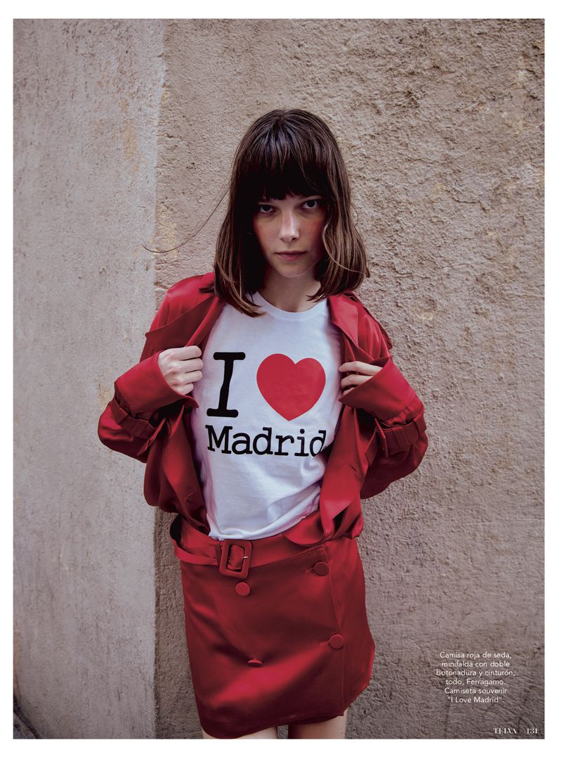 A brunette woman in a red skirt and jacket poses against a textured wall, wearing a t-shirt that says, 'I Heart Madrid'.