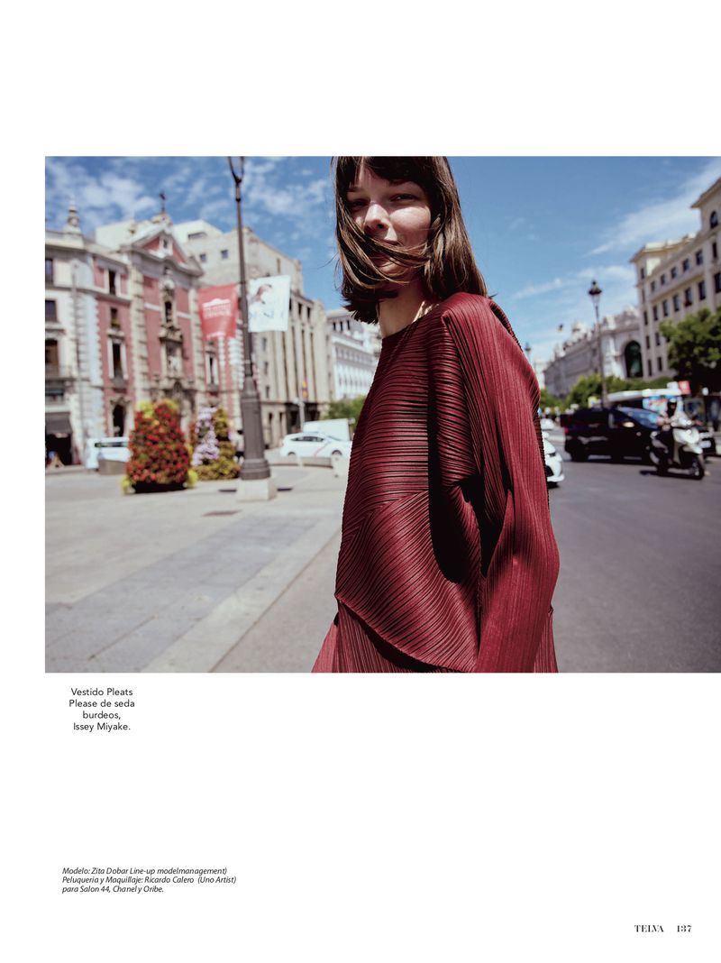 Fashion editorial shot of a woman in a pleated red dress standing on a city street with historic buildings and cars in the background.