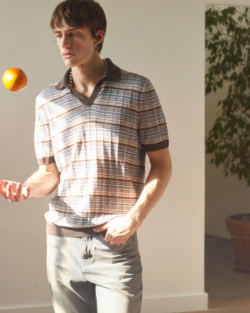 A young adult male stands indoors in a stylish polo shirt and light jeans, tossing a bright orange fruit in the air in soft, diffused light.