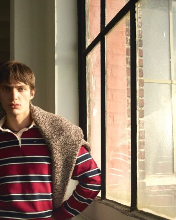 Man in striped rugby shirt leans against window with sunlight, wearing a sweater over shoulders. Natural light, indoor setting.