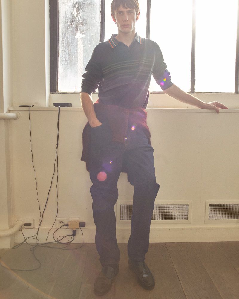 Full-length studio shot of a young adult male fashion model in a dark polo shirt and blue pants, leaning against a window.