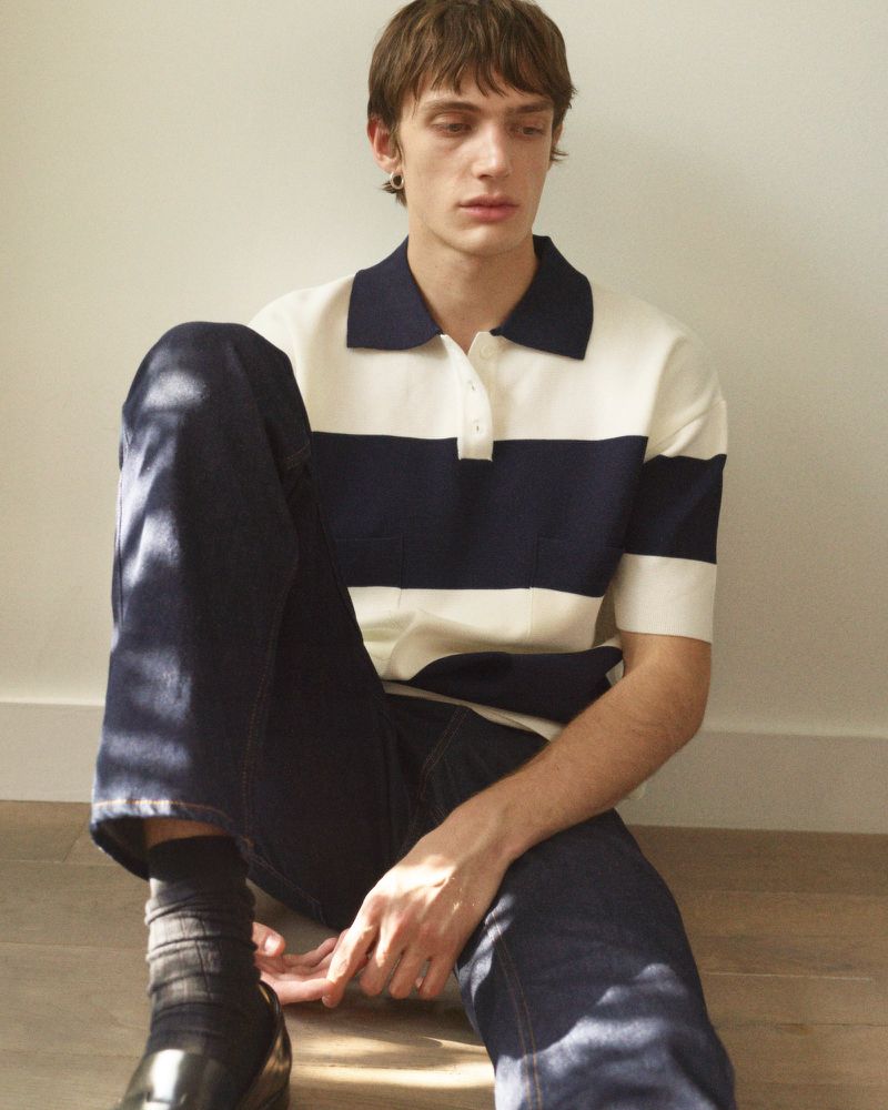 A slender man wearing a rugby shirt sits on a light wooden floor in a relaxed pose, looking off-camera in a soft, diffused light.