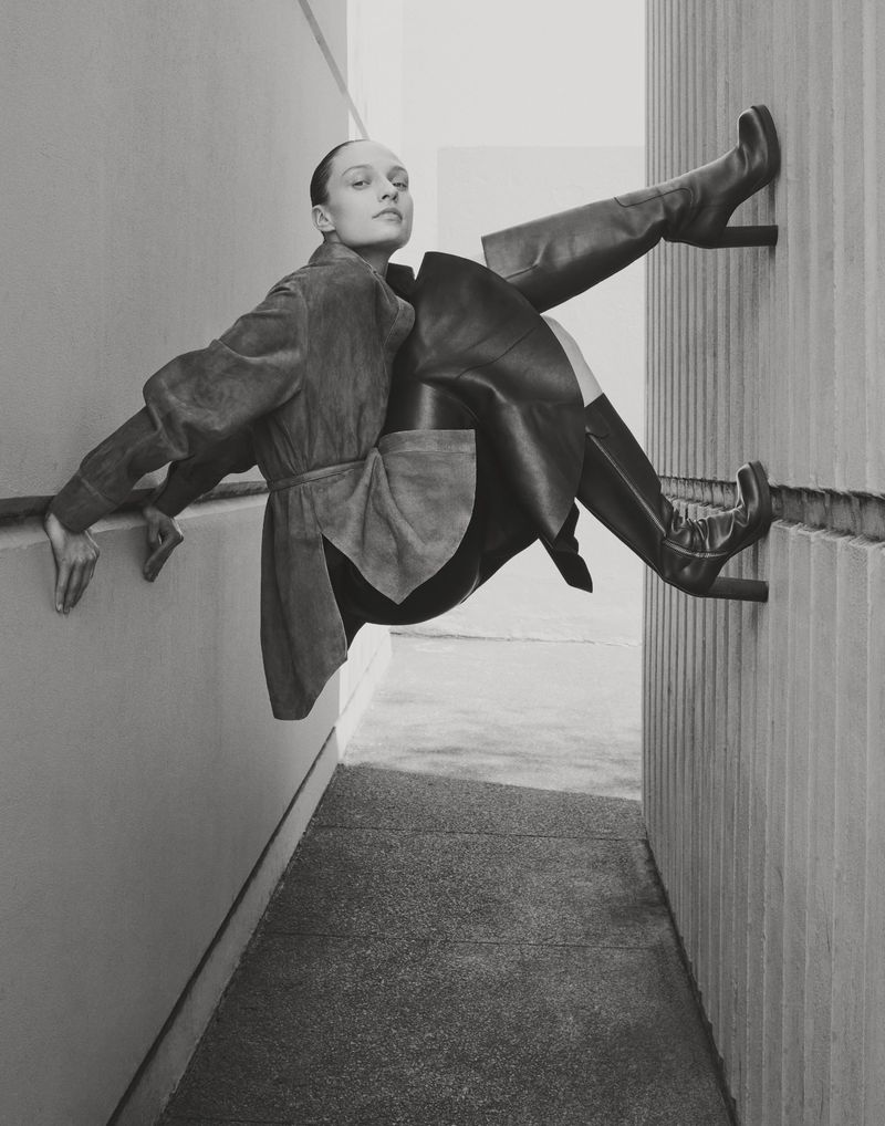 Monochrome fashion shot: a female model in a coat and high boots poses in an alleyway, emphasizing lines and geometric shapes.