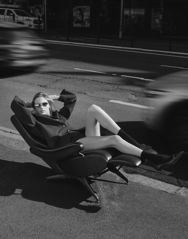 Monochrome fashion shot of a woman in sunglasses relaxing on an indoor/outdoor chair placed on an asphalt road with cars in background.