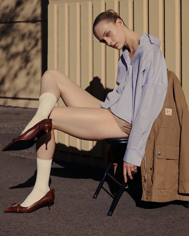 A young adult female is seated, modeling a striped shirt, brown shoes, and tan shorts, accompanied by a tan jacket in an outdoor setting.