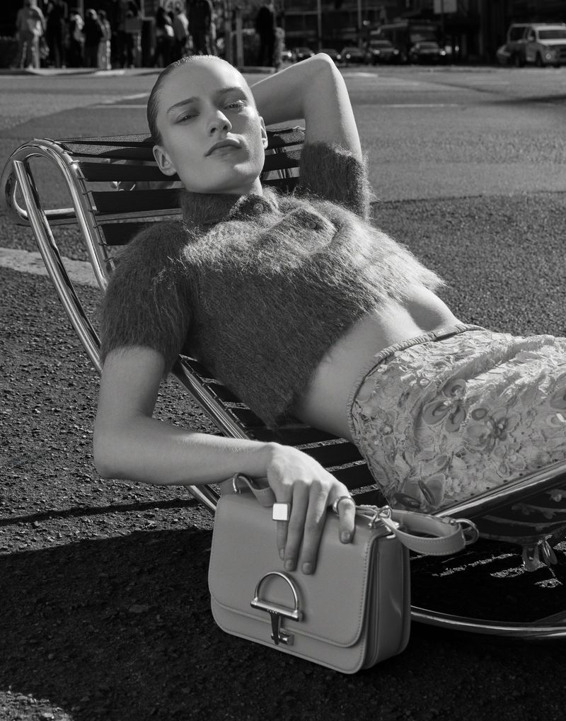 Monochrome outdoor fashion shot of a woman lying in a chair with a purse in her lap, with city street background, shallow depth of field.