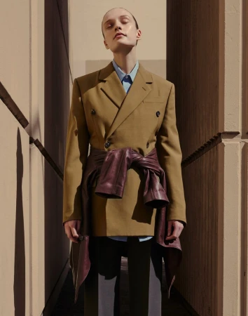 A woman stands in a beige blazer with a purple jacket tied around her waist in front of a beige wall and architecture.