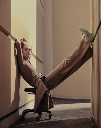 Woman in brown suit rests on office chair against wall. Fashion shot with studio lighting and neutral color palette with long exposure.