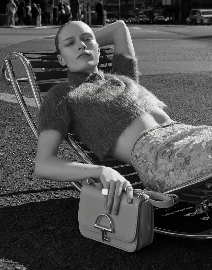 Model reclining on a chrome lounge chair on an asphalt street wearing a fuzzy crop sweater and resting her hand on a leather bag.