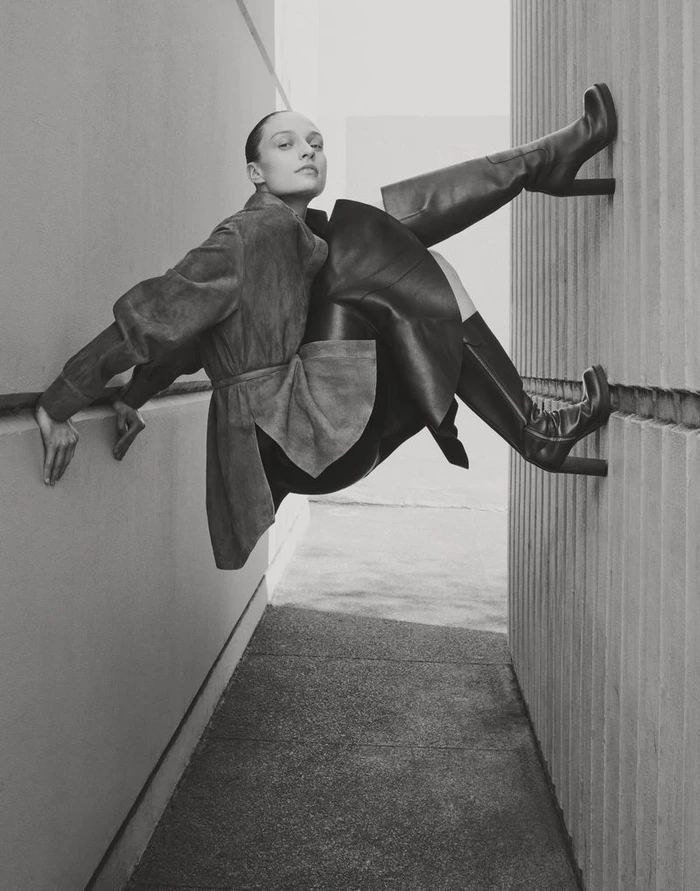 Model Katie Robinson poses suspended between two concrete walls wearing a belted jacket and high-heel knee-high boots.