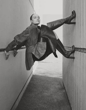 Model Katie Robinson poses suspended between two concrete walls wearing a belted jacket and high-heel knee-high boots.