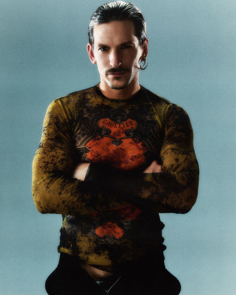 Portrait of a man with a mustache and ear piercings, wearing a patterned long-sleeved shirt against a pale blue background in a studio setting.
