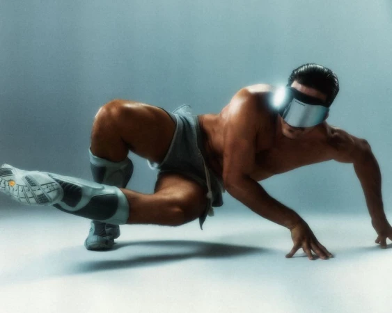 Athletic adult male posing on his hands and foot, wearing futuristic visor, shorts and knee pads against a pale blue backdrop.