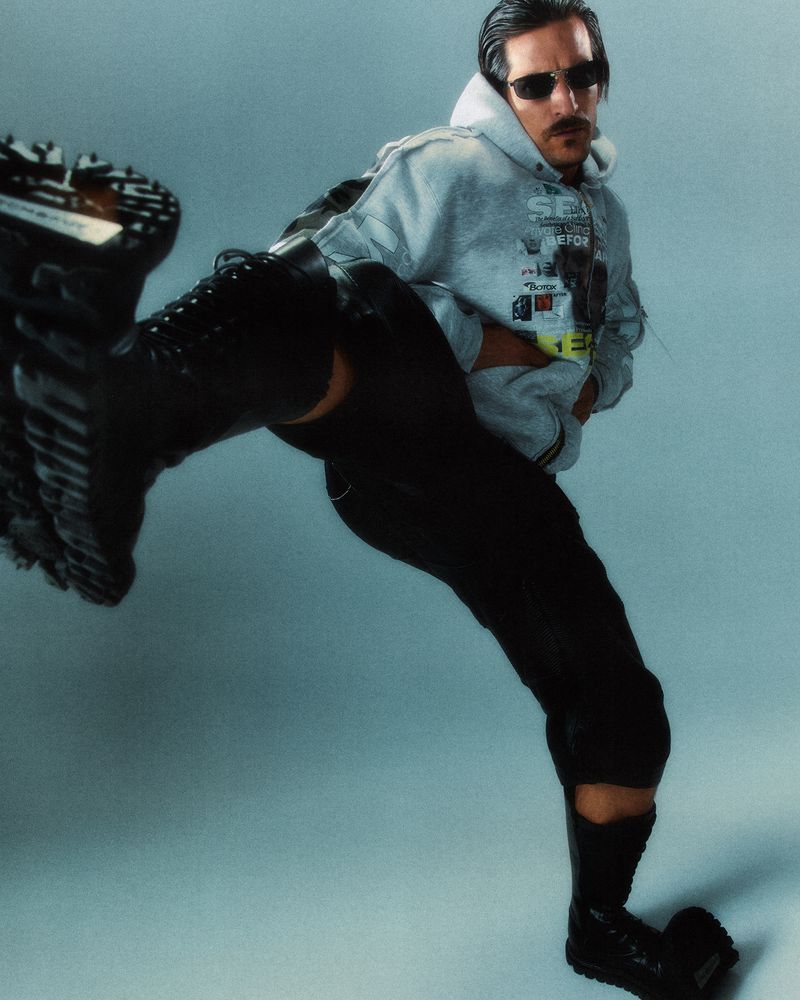 Person in a grey hoodie, black pants and boots strikes a dramatic pose against a light blue background, showcasing street fashion.