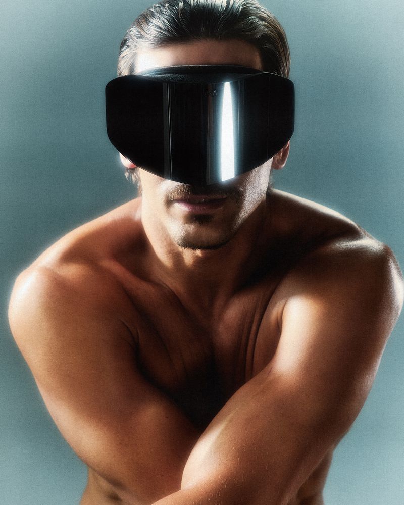 Man with a muscular physique wears futuristic black eyewear that covers his eyes and nose in a close-up studio portrait.
