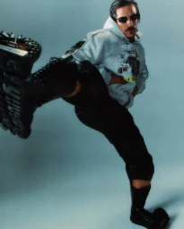 Model Jarrod Scott performing a dynamic high kick wearing a grey hooded sweatshirt, black combat boots, and dark sunglasses.