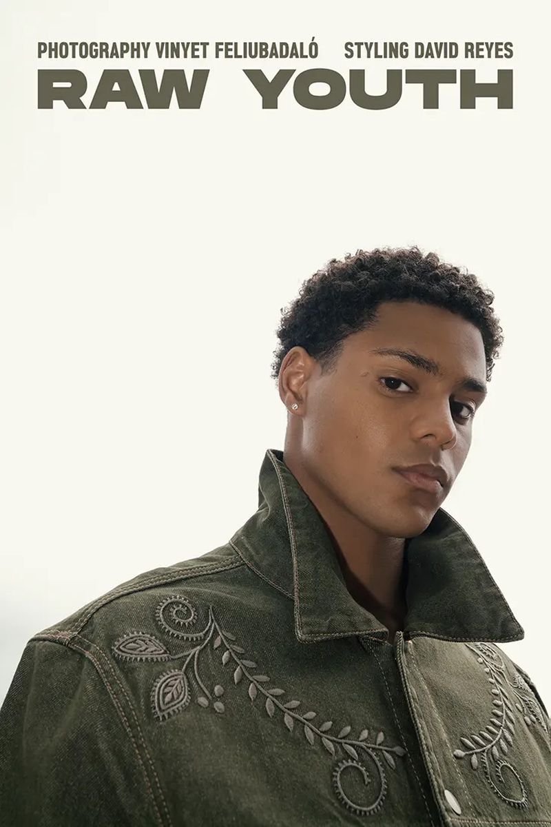 Portrait of a young adult male with curly hair wearing a green denim jacket, set against a neutral background in a fashion editorial.