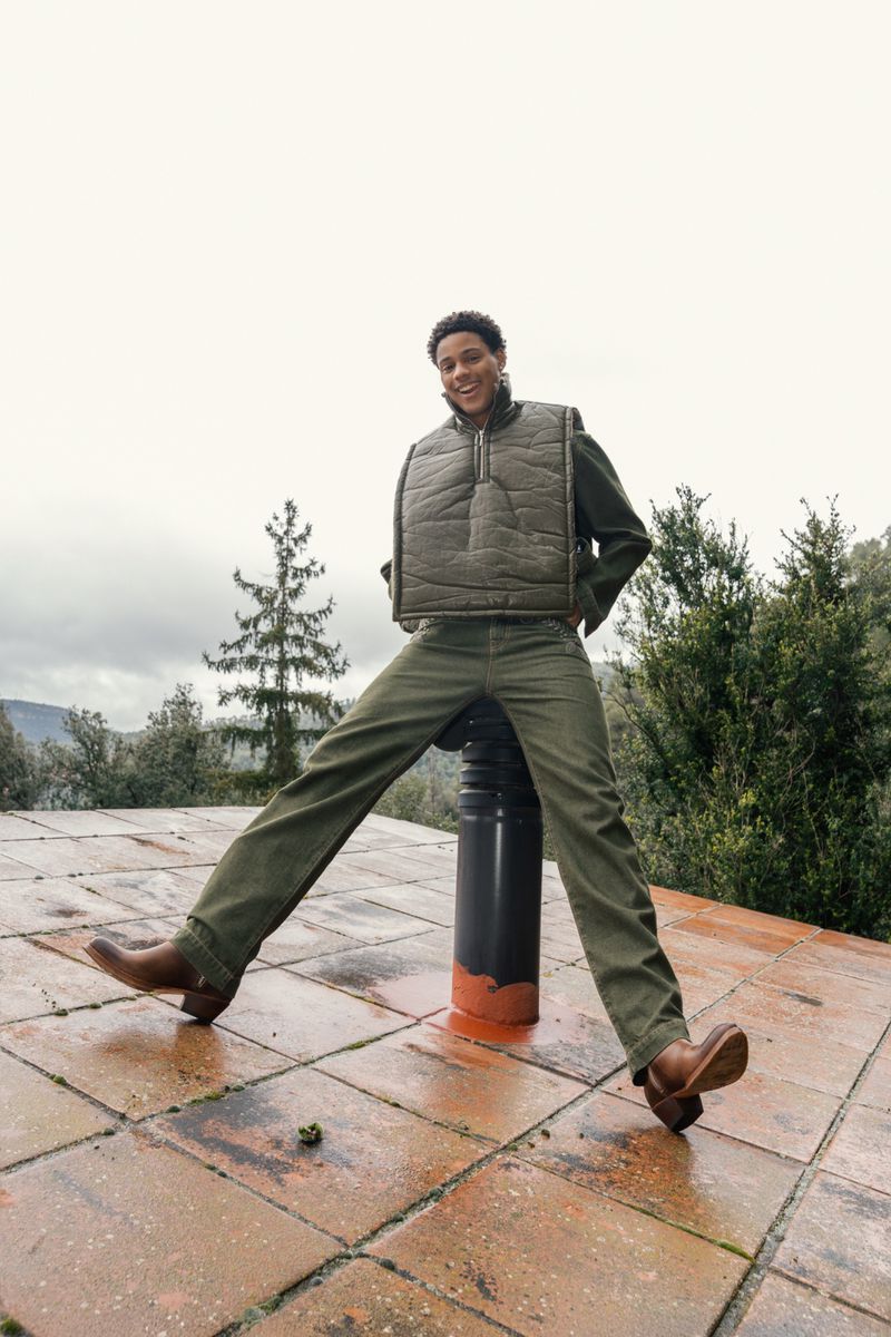 A young adult poses outside wearing a puffer vest, jeans, and cowboy boots, with a forest and cloudy sky visible in the background.